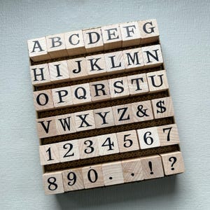 May include: A set of square wooden stamps arranged in a rectangular box. The stamps feature black printed characters including the uppercase alphabet from A to Z, numbers 0 through 9, an ampersand, a dollar sign, a period, an exclamation point, and a question mark.