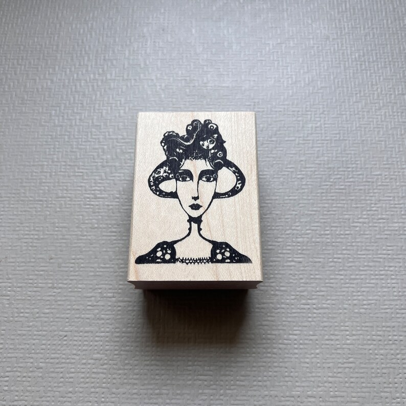 May include: A black and white rubber stamp featuring a woman with a stylized, intricate hairstyle. The woman is wearing a decorative, patterned dress.