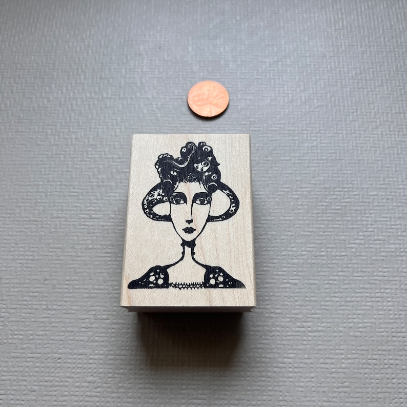 May include: A wooden stamp with a black and white illustration of a woman with an octopus for hair. The woman is wearing a patterned dress.