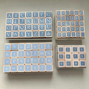 May include: Four sets of wooden alphabet and number stamps. The stamps have a light blue background with white letters and numbers. The sets include uppercase and lowercase letters, numbers, and punctuation marks.