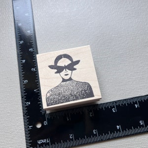 May include: A wooden stamp with a black and white illustration of a person with butterfly wings over their eyes. The person is wearing a patterned shirt.