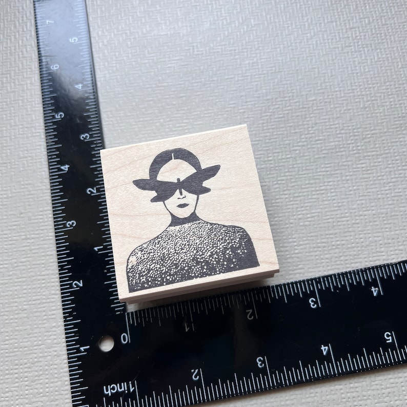 May include: A wooden stamp with a black and white illustration of a person with butterfly wings over their eyes. The person is wearing a patterned shirt.