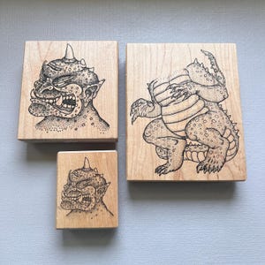 May include: Set of three wooden stamps featuring detailed black and white illustrations of fantastical creatures. The designs include a horned monster face and a squatting, reptilian-like figure. The stamps are on light-colored wooden blocks.