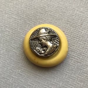 May include: A silver button with a raised image of a person wearing a hat. The button is set in a yellow plastic ring.