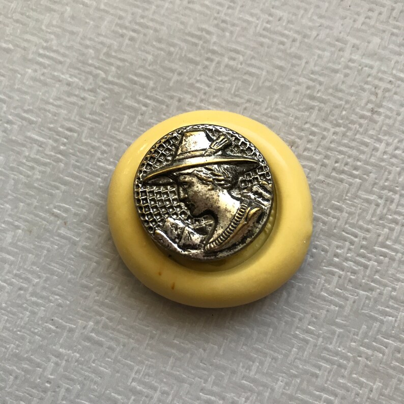 May include: A silver button with a raised image of a person wearing a hat. The button is set in a yellow plastic ring.
