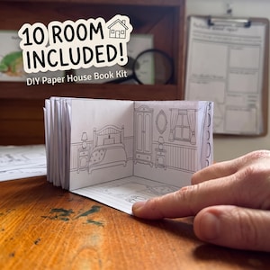 May include: A DIY paper house book kit featuring 10 rooms. The open book displays a black and white line drawing of a bedroom with a bed, wardrobe, mirror, and window. The text 10 ROOM INCLUDED! DIY Paper House Book Kit is visible above.