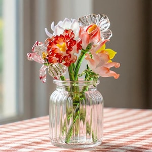 May include: A clear, ribbed glass vase containing a bouquet of handcrafted glass flowers. The arrangement features various blooms in shades of red, orange, yellow, and white, each with green glass stems visible through the transparent glass container.