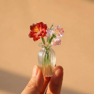 May include: A miniature glass vase with vertical ribbing containing two delicate glass flowers. One flower is deep red with a yellow center, and the other is translucent pink with white edges. Both flowers have green glass stems visible inside.