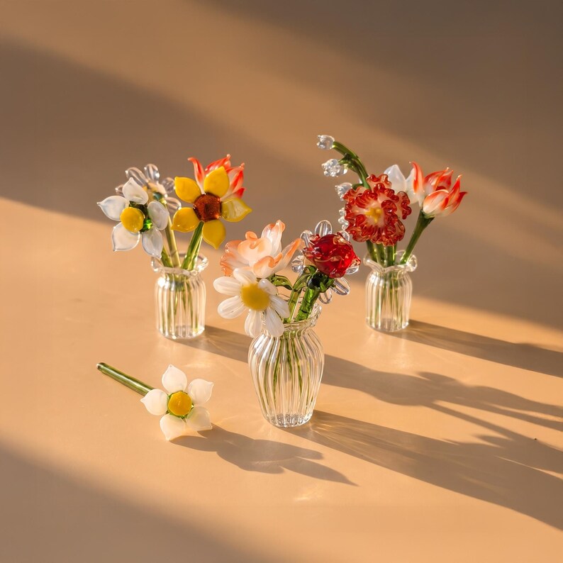May include: Three miniature clear ribbed glass vases each containing small handcrafted glass flowers in vibrant shades of red, orange, yellow, and white, with one additional white glass flower lying separately on the surface.