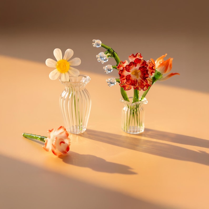 May include: Two miniature clear glass vases with vertical ribbing. One vase holds a white daisy with a yellow center. The other vase contains a cluster of lily of the valley and red-tipped flowers. A single red-tipped flower lies on the surface.