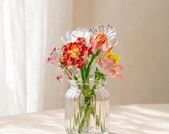 May include: A clear, ribbed glass vase containing a bouquet of handcrafted glass flowers. The arrangement features various blossoms in shades of red, pink, white, and yellow, each with green glass stems visible through the transparent, fluted vessel.