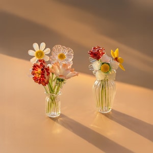 May include: Two miniature glass vases containing small, handcrafted glass flower arrangements. The left vase holds white, red, and peach-colored blossoms. The right vase features a red rose, white buds, and a yellow flower, all with green stems.