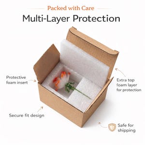 May include: A brown cardboard box containing a small glass flower with white and orange petals and a green stem. The box features white foam inserts for protection, with text labels reading Packed with Care, Multi-Layer Protection, Protective foam insert, Extra top foam layer for protection, Secure fit design, and Safe for shipping.