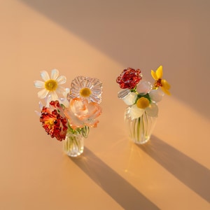 May include: Two miniature glass vases containing small, handcrafted glass flower arrangements. The left vase holds white, orange, and deep red blossoms. The right vase features a red rose, white petals, and a bright yellow flower, all made of glass.