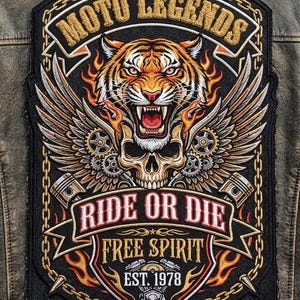 Pode incluir: Um grande emblema bordado numa jaqueta de couro castanha com uma cabeça de tigre sobre um crânio com asas, engrenagens e pistões. O texto diz MOTO LEGENDS, RIDE OR DIE, FREE SPIRIT e EST. 1978, emoldurado por uma corrente dourada.