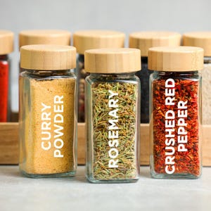 Puede incluir: Una colección de botes de especias de vidrio con tapas de madera. Los botes contienen curry en polvo, romero y pimiento rojo triturado. Cada bote tiene una etiqueta blanca con el nombre de la especia. Los botes están colocados en un estante de madera.
