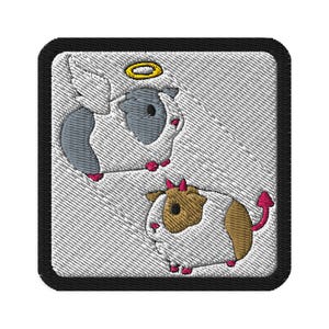 Może przedstawiać: Haftowana naszywka z czarną obwódką, przedstawiająca dwie świnki morskie. Jedna to anioł ze skrzydłami i aureolą, druga to diabeł z rogami i ogonem. Na teksturowanym białym tle.