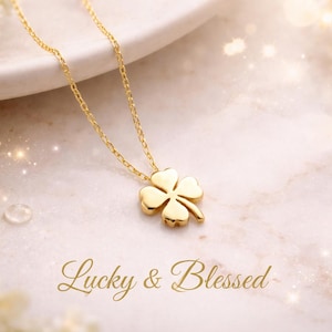 Op de afbeelding: Een goudkleurige klaverblad hanger ketting. De bedel hangt aan een delicate ketting. De woorden "Lucky & Blessed" staan in gouden letters eronder. De achtergrond is zacht, wazig, lichtroze en wit.