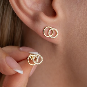 May include: A pair of gold stud earrings, each featuring two interlocking circles. One circle is a plain polished gold band, while the other is a gold band encrusted with small, clear, sparkling stones. The earrings have a secure post backing.