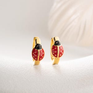 Puede incluir: Un par de pendientes de aro dorados con un dije de mariquita. Cada mariquita tiene un dorso de esmalte rojo brillante con pequeños puntos dorados en relieve y una cabeza de esmalte negro. Los aros son de metal dorado pulido.