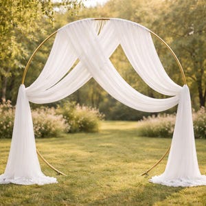 Può includere: Un arco nuziale in metallo dorato con una struttura circolare, drappeggiato con tessuto bianco fluente. L'arco è posizionato all'aperto su un prato, creando un'elegante cornice per cerimonie ed eventi.
