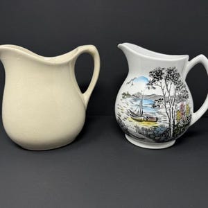May include: Two ceramic pitchers. One is a solid cream color, the other is white with a landscape scene featuring a sailboat, trees, and colorful flowers. Both pitchers have a curved spout and a handle.