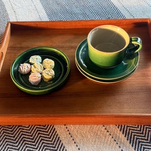 Könnte beinhalten: Ein Holztablett mit einer grünen Tasse und Untertasse sowie einer kleinen grünen Schale mit bunten Süßigkeiten. Die Tasse hat eine gelbe Innenseite und einen grünen Henkel. Die Süßigkeiten sind pastellfarben.