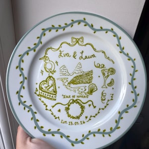 May include: A white ceramic plate with a light blue scalloped border featuring green leaf accents. The centre displays green line art including the text 'Eva & Laura', 'ISLE OF HARRIS', and 'EST. 23.07.26', alongside illustrations of a ring, a heart cake, a landscape, a cocktail, and a wreath.