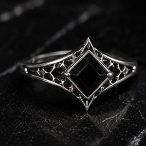 May include: A silver ring featuring a square-cut black gemstone set in a diamond-shaped frame. The band displays an intricate, openwork lattice design that extends from the central setting, showcasing a polished metallic finish throughout.