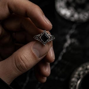 May include: A silver ring featuring a central square-cut black stone set within a pointed, diamond-shaped frame. The band displays an intricate, openwork filigree pattern that extends from the central setting, creating a detailed metalwork design.