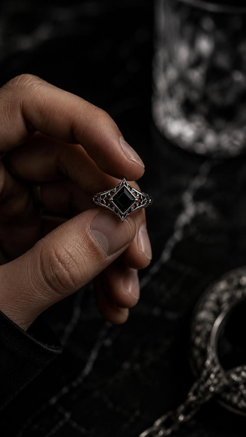 May include: A silver ring featuring a central square-cut black stone set within a pointed, diamond-shaped frame. The band displays an intricate, openwork filigree pattern that extends from the central setting, creating a detailed metalwork design.