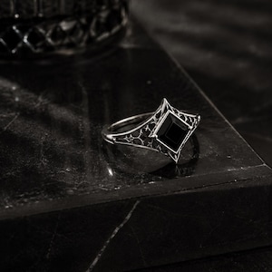 May include: A silver-toned ring featuring a central square-cut black gemstone set in a pointed, diamond-shaped frame. The band displays intricate, openwork filigree detailing that extends from the setting along the sides of the ring.