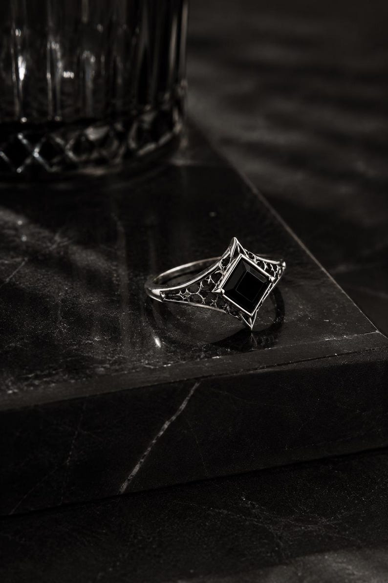 May include: A silver-toned ring featuring a central square-cut black gemstone set in a pointed, diamond-shaped frame. The band displays intricate, openwork filigree detailing that extends from the setting along the sides of the ring.