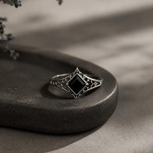 May include: A silver ring featuring a square-cut black gemstone set in a diamond-shaped frame. The band displays an intricate, openwork filigree pattern that extends from the central setting, creating a detailed metalwork design throughout the piece.