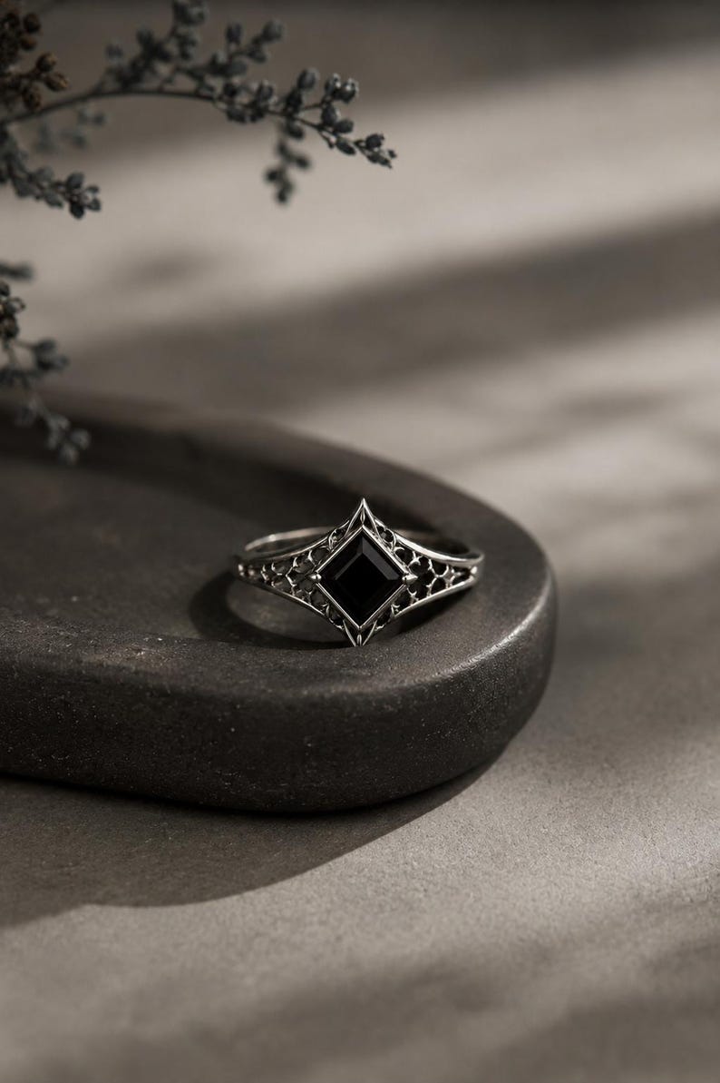 May include: A silver ring featuring a square-cut black gemstone set in a diamond-shaped frame. The band displays an intricate, openwork filigree pattern that extends from the central setting, creating a detailed metalwork design throughout the piece.