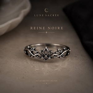 May include: A 925 sterling silver ring featuring an intricate, oxidized filigree design with a central fleur-de-lis motif. The text LUNE SACRÉE, REINE NOIRE, and 925 STERLING SILVER is visible above and below the ring in a dark, elegant font.