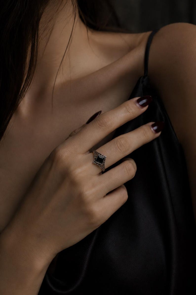 May include: A silver-toned ring featuring a square-cut black gemstone set in a detailed, openwork filigree band. The metalwork displays intricate, lace-like patterns surrounding the central stone, creating a delicate and ornate appearance.