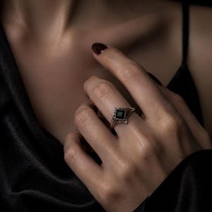 May include: A silver ring featuring a square-cut black gemstone set in a pointed, openwork metal band with intricate filigree detailing. The band has a geometric, angular design that tapers toward the back, showcasing a dark, polished finish.