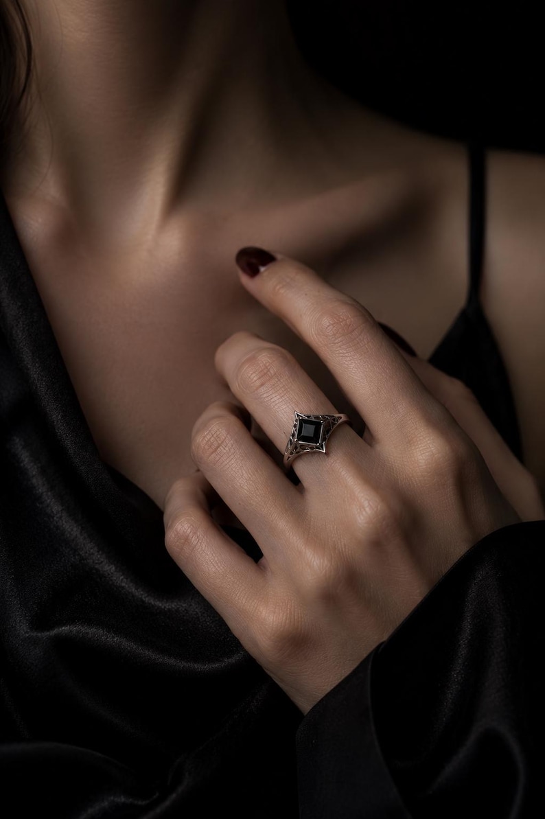 May include: A silver ring featuring a square-cut black gemstone set in a pointed, openwork metal band with intricate filigree detailing. The band has a geometric, angular design that tapers toward the back, showcasing a dark, polished finish.