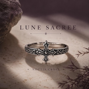 May include: A silver ring with a cross design. The ring features an ornate cross in the center and decorative patterns around the band. The words "LUNE SACREE" and "NOCTIS CROIX" are printed on the image.