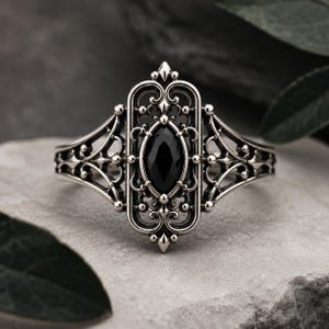May include: A silver-toned ring featuring an elongated, rectangular filigree setting with intricate scrollwork and bead accents. A central, marquise-cut black gemstone is securely mounted within the detailed metal frame, creating a balanced design.