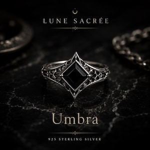 May include: A 925 sterling silver ring featuring a central square-cut black stone set within an intricate, openwork metal frame. The text LUNE SACRÉE, Umbra, and 925 STERLING SILVER is visible. The design includes pointed edges and filigree details.