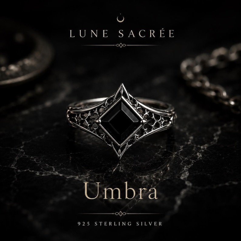 May include: A 925 sterling silver ring featuring a central square-cut black stone set within an intricate, openwork metal frame. The text LUNE SACR&Eacute;E, Umbra, and 925 STERLING SILVER is visible. The design includes pointed edges and filigree details.