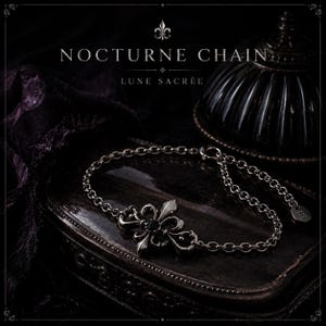 Op de afbeelding: Een zilveren armband met een fleur-de-lis-ontwerp en zwarte details. De armband wordt gepresenteerd op een donker, sierlijk oppervlak. De tekst "NOCTURNE CHAIN" en "LUNE SACRÉE" zijn zichtbaar.