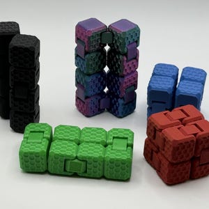 May include: A selection of colourful fidget toys in various shapes. The toys are in shades of black, green, blue, red, and a rainbow gradient. Each toy is made of connected blocks with a textured surface.