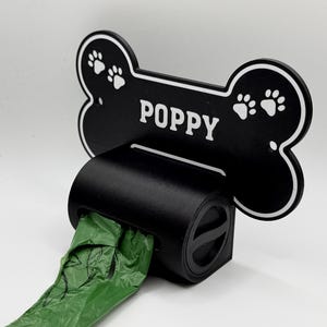 Peut inclure: Distributeur de sacs à déjections canines noir en forme d'os, avec le nom "POPPY" en blanc. L'os comporte des empreintes de pattes blanches. Un rouleau de sacs verts est visible.