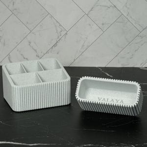 May include: Two light gray organizers. One is a rectangular container with six compartments. The other is a smaller rectangular tray with the word "VALAYA" printed on the inside. Both have vertical ribbed detailing.