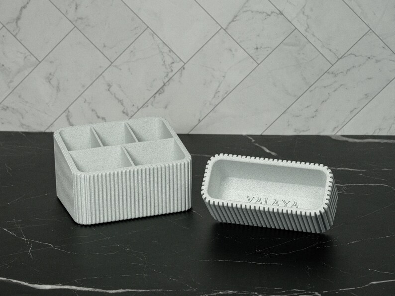May include: Two light gray organizers. One is a rectangular container with six compartments. The other is a smaller rectangular tray with the word "VALAYA" printed on the inside. Both have vertical ribbed detailing.