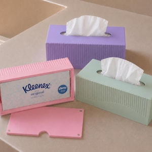 May include: Three pastel-coloured tissue boxes: lavender, mint green, and pink. The pink box is open, showing a Kleenex box. Each box is rectangular with a vertical ribbed design and an oval opening for tissue dispensing.