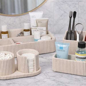 May include: Beige ribbed bathroom organizers holding toiletries, toothbrushes, toothpaste, and mouthwash. The set includes a tray with containers for cotton swabs and pads, and a larger tray with skincare products.